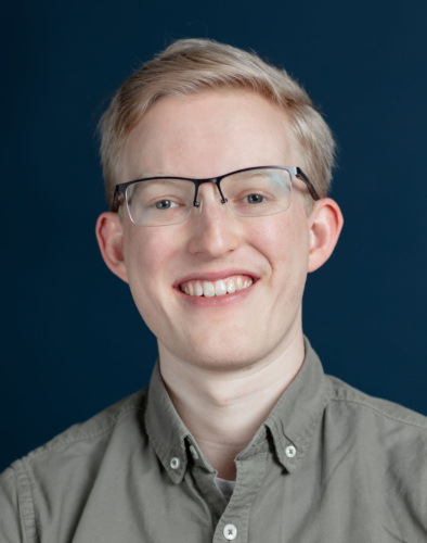 A person with short blond hair and glasses smiles at the camera, wearing a gray button-up shirt against a dark blue background.